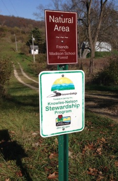 Two signs on a post that mark the drive into the Friends Property. One says "Natural Area - Friends of the Madison School Forest" and the other says "Stewardship funded in part by the Knowles-Nelson Stewardship Program, Wisconsin Department of Natural Resources"